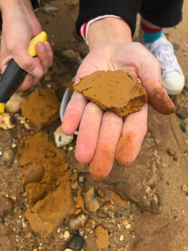 Picture shows a ladies hand outstretched with a piece of ochre freshly dug up from the beach in the background of the shot.